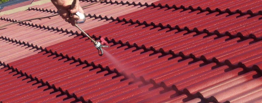 Hand holding a spray gun applying a vibrant red coating to faded terracotta roof tiles.